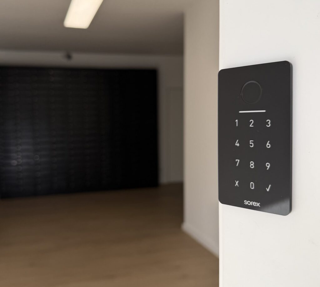 Modernes Zutrittssystem mit Zahlencode-Tastatur im Smarvo Gebäude, montiert an einer weißen Wand mit Holzfußboden und Briefkastenanlage im Hintergrund.