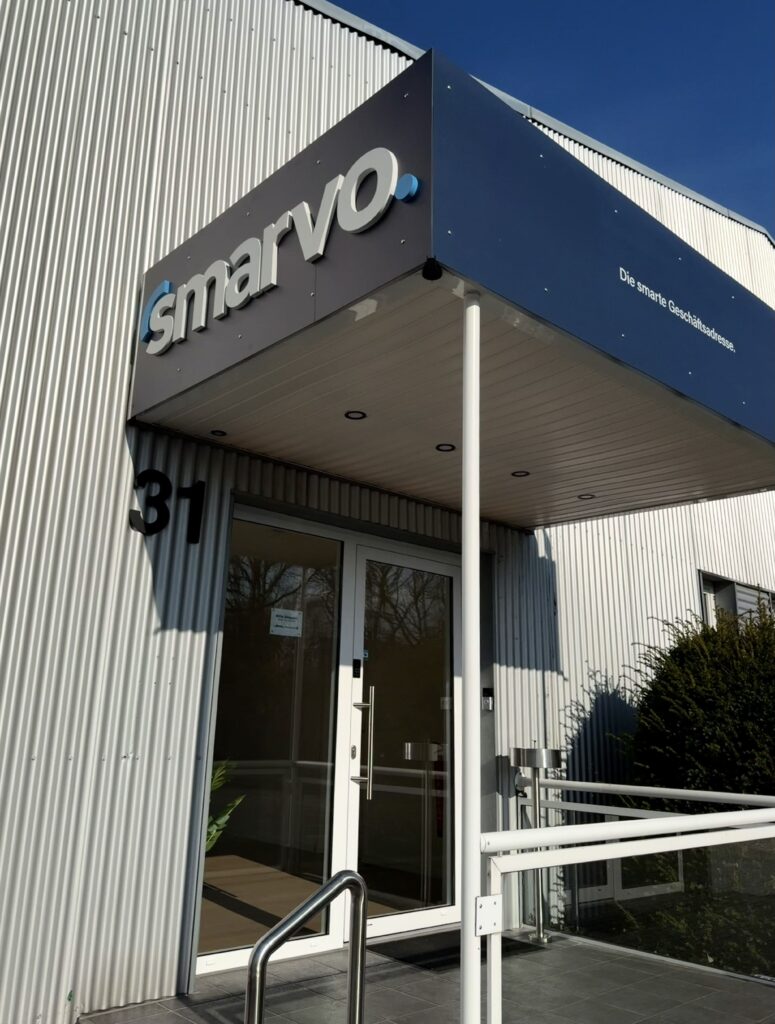 Außenfassade des Smarvo-Gebäudes mit blauem Vordach, 3D-Logo und dem Slogan „Die smarte Geschäftsadresse“. Der Eingang aus Glas hat die Hausnummer 31.