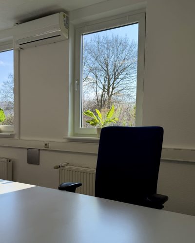 Moderner Büroarbeitsplatz mit dunkelblauem ergonomischen Stuhl, weißem Schreibtisch, wandmontierter Klimaanlage und großem Fenster mit Blick auf den Parkplatz im grünen.
