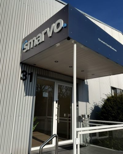 Außenfassade des Smarvo-Gebäudes mit blauem Vordach, 3D-Logo und dem Slogan „Die smarte Geschäftsadresse“. Der Eingang aus Glas hat die Hausnummer 31.