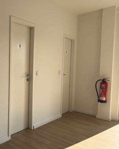 Heller Flur mit Boden in Holzoptik, zwei weißen Türen für getrennte Damen- und Herren-WCs sowie einem rot montierten Feuerlöscher an der Wand.